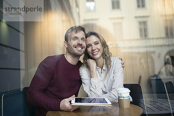 Smiling boyfriend with arm around girlfriend and holding tablet PC in cafe