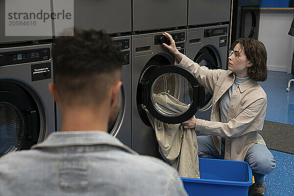 Young woman operating washing machine near boyfriend at laundromat