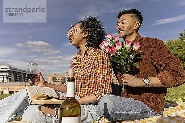 Smiling man holding bunch of flowers and covering girlfriend's eyes at park