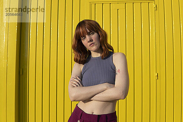 Confident redhead woman with arms crossed standing in front of yellow wall on sunny day