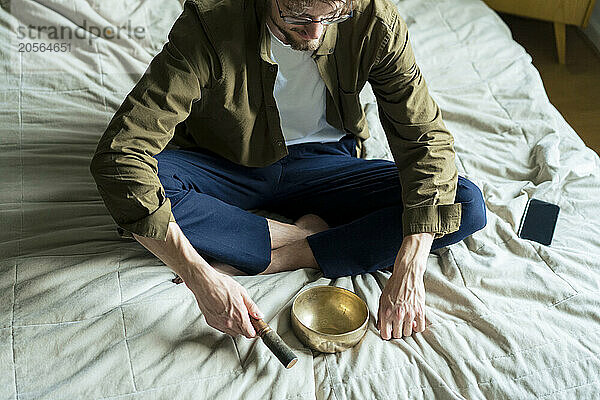 Young man meditating with singing bowl on bed at home