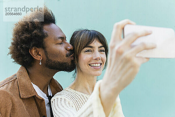 Smiling woman taking selfie with boyfriend kissing her in front of blue wall