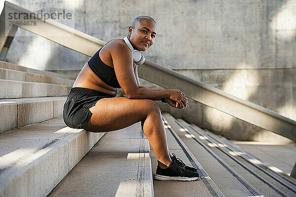 Smiling muscular woman with headphones sitting on stairs