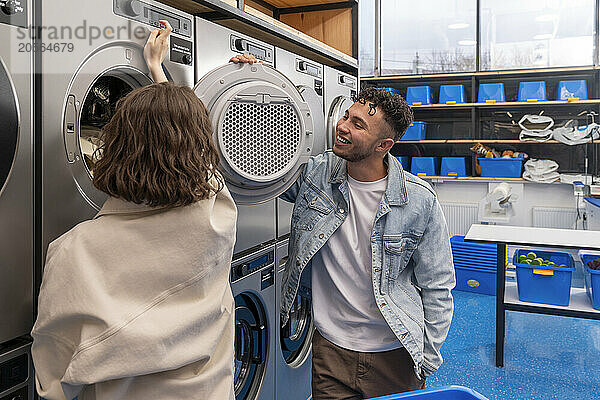 Smiling young man looking at girlfriend operating dryer in laundromat