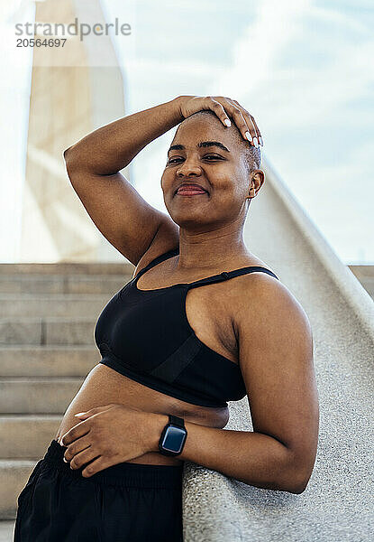 Smiling muscular woman touching head leaning on railing