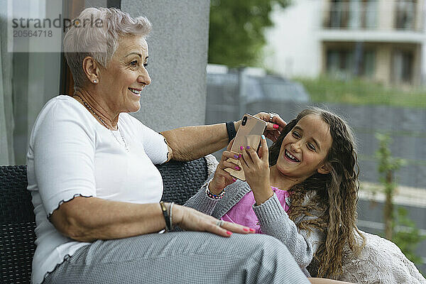 Happy girl photographing grandmother through smart phone at patio