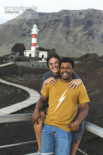 Happy young woman with arm around boyfriend posing in front of Punta Teno Lighthouse