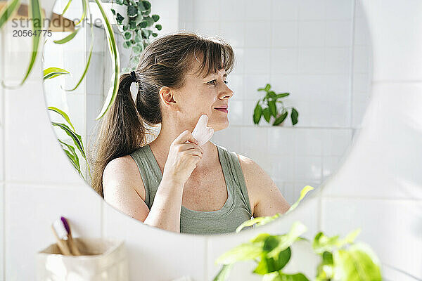 Smiling woman massaging face with gua sha stone in bathroom