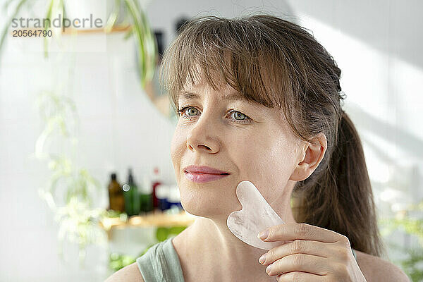 Mature woman massaging face with gua sha stone in bathroom