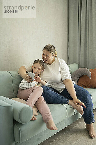 Mother using smart phone with daughter sitting on sofa at home