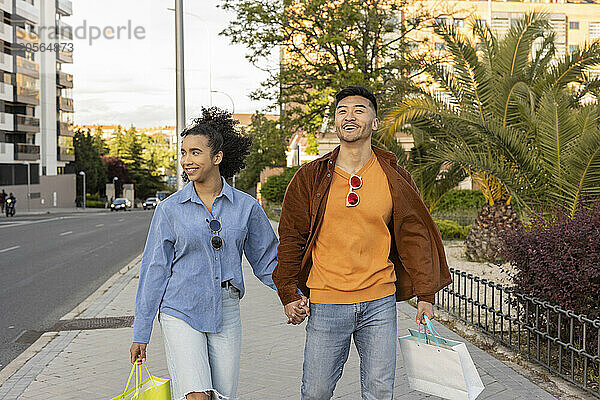 Happy boyfriend and girlfriend carrying shopping bags and walking on footpath