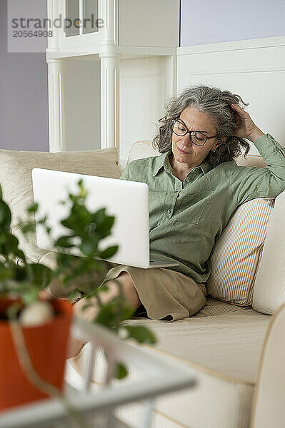 Tired businesswoman with hand in hair and sitting with laptop on sofa at home