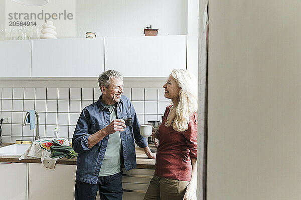 Smiling retired senior couple with coffee cups talking in kitchen at home