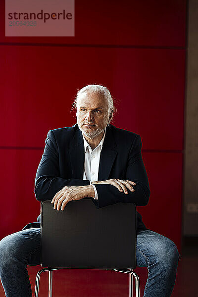 Confident businessman sitting on chair in front of red wall at office