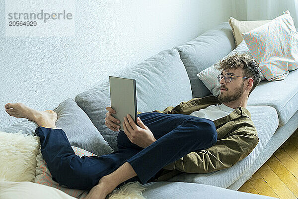 Flexible young man using tablet PC lying on sofa at home