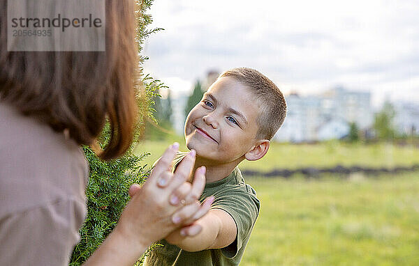 The boy looks from behind a tree at his mother  smiles and holds her hand.