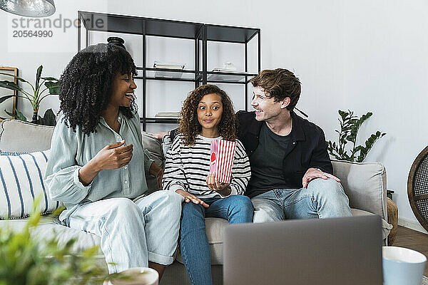 Parents and daughter sitting on sofa eating popcorn watching movie on laptop