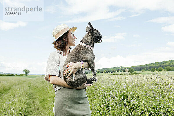 Smiling woman standing and carrying pet french bulldog in field