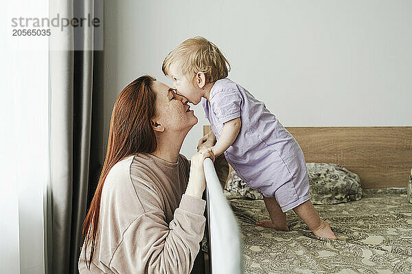 Cute baby girl biting redhead mother's nose in bedroom