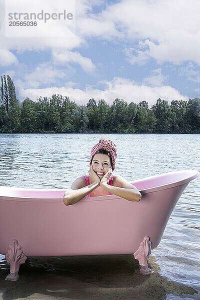 Smiling girl sitting inside pink bathtub with towel wrapped on head in lake