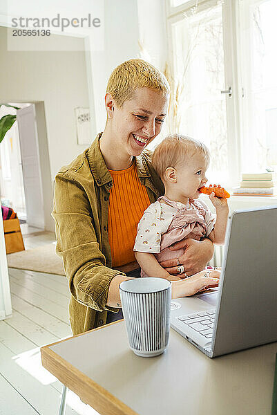 Smiling working mother using laptop sitting with daughter eating carrot at home