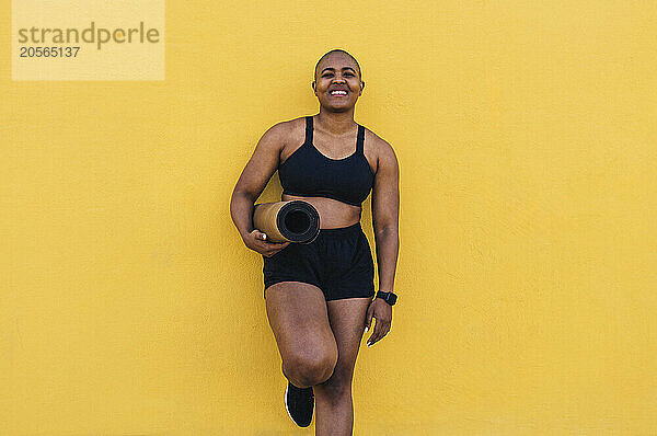 Happy woman holding exercise mat and leaning on yellow wall