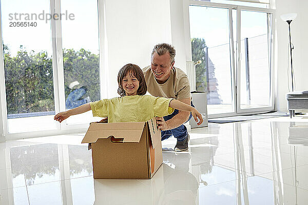 Happy father pushing son with arms outstretched in cardboard box at new empty house