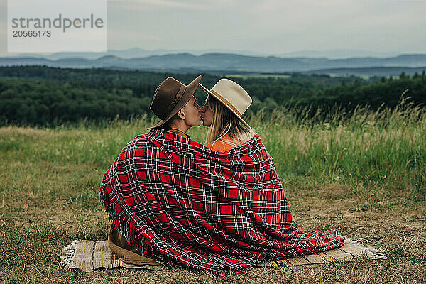 Romantic young couple in plaid shawl sitting and kissing at meadow in mountain of Poland