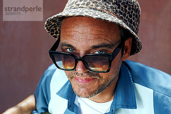 Smiling man wearing sunglasses and leopard print hat in front of wall