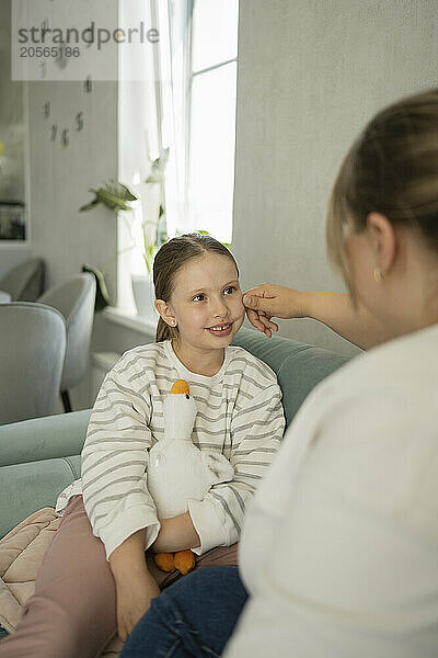 Mother touching cheek of daughter sitting on sofa at home