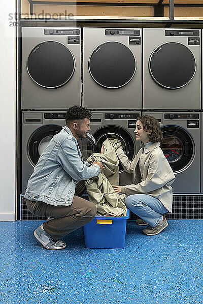 Young couple removing sheet from washing machine squatting in laundromat