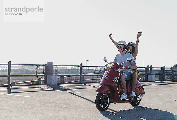 Happy young woman with arms raised sitting on motor scooter with boyfriend at parking lot