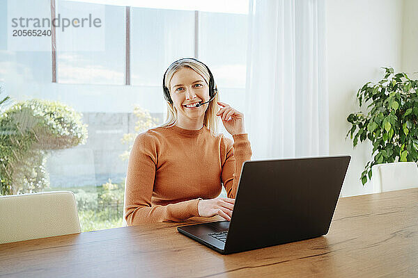 Smiling young businesswoman with headset using laptop at table working from home