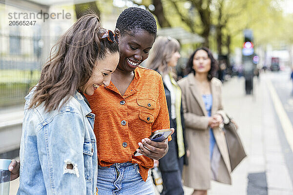 Smiling woman sharing smart phone with friend at sidewalk