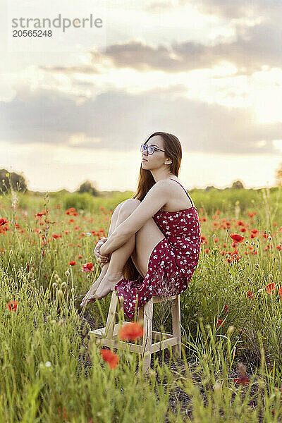 Contemplative young woman sitting on stool in poppy field at sunset