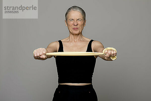 Senior woman exercising with resistance band grimacing against gray background