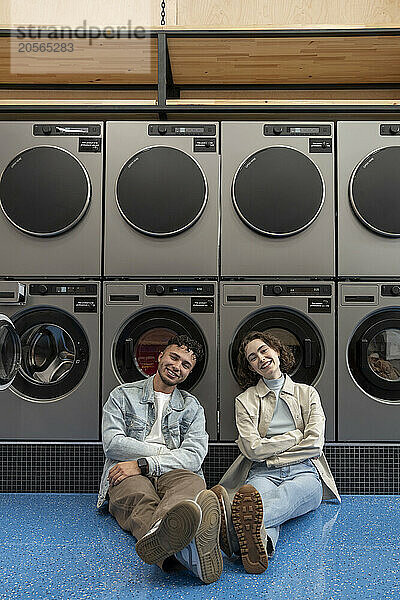 Smiling young couple leaning on washing machines sitting together in laundromat