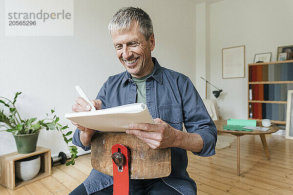 Happy senior man sitting on chair writing in diary at home