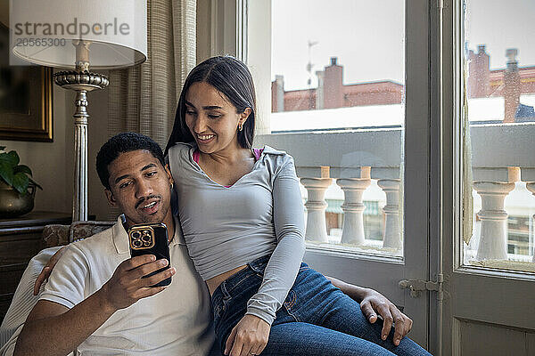 Young man sharing smart phone with girlfriend sitting at home