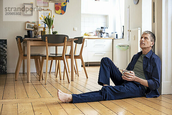 Thoughtful senior man with coffee cup sitting on hardwood floor at home