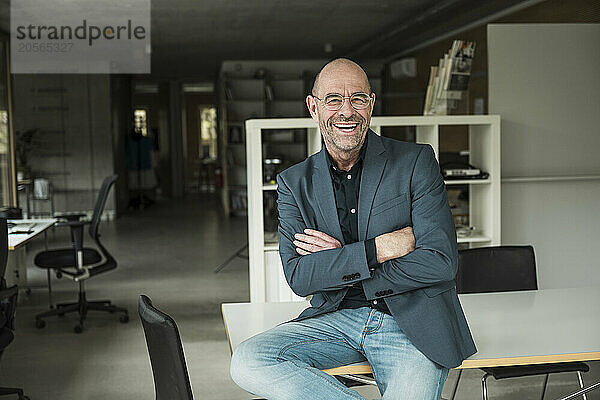 Happy shaved head businessman with arms crossed sitting on desk in office