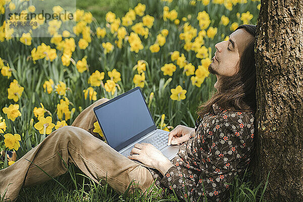 Tired man with laptop leaning on tree trunk sleeping near daffodil flowers