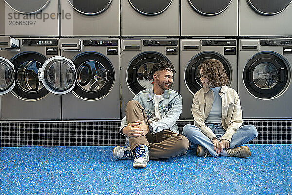 Smiling couple sitting together leaning on washing machines in laundromat