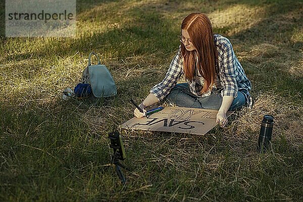 Redhead teenage girl making protest banner and vlogging through smart phone sitting on grass at park