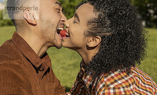 Happy couple eating strawberry from mouth to mouth at park