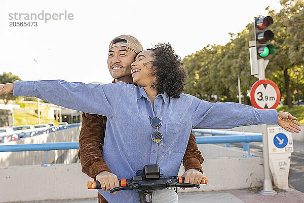 Happy woman with arms outstretched and standing with boyfriend on electric push scooter at park