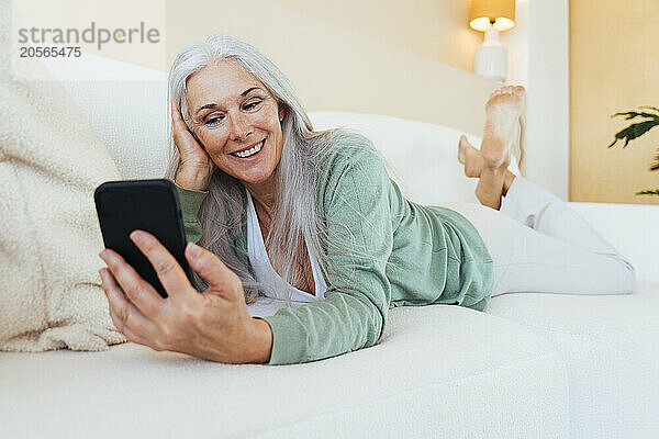 Happy mature woman lying on sofa and using smart phone at home