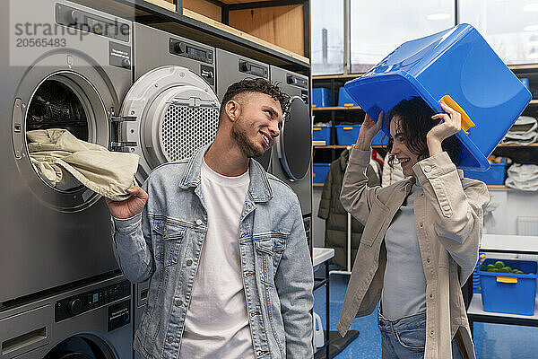 Smiling man looking at playful girlfriend with blue basket in laundromat