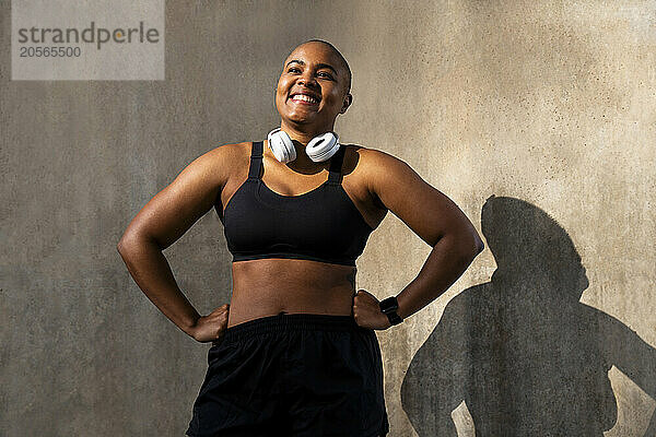 Smiling sporty woman with arms akimbo in front of wall