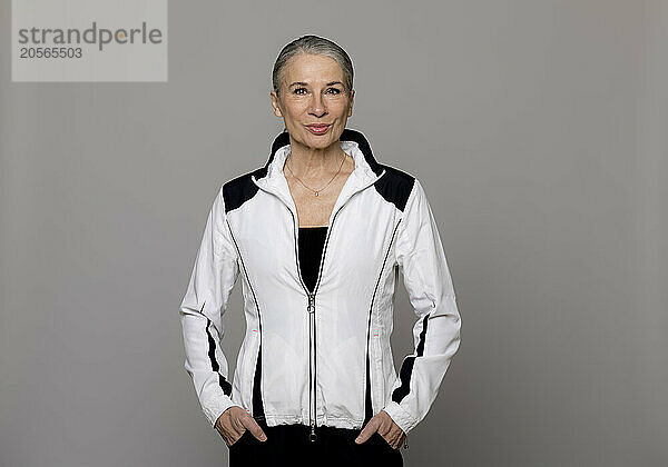 Smiling senior woman standing with hands in pockets against gray background